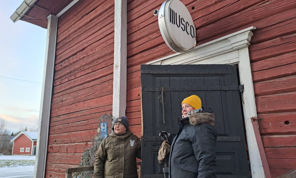 Kaksi naista seisoo vanhan punaisen rakennuksen portailla oven edessä. Oven päällä pyöreä kyltti, jossa lukee museo. Toisella naisista on keltainen pipo päässä.