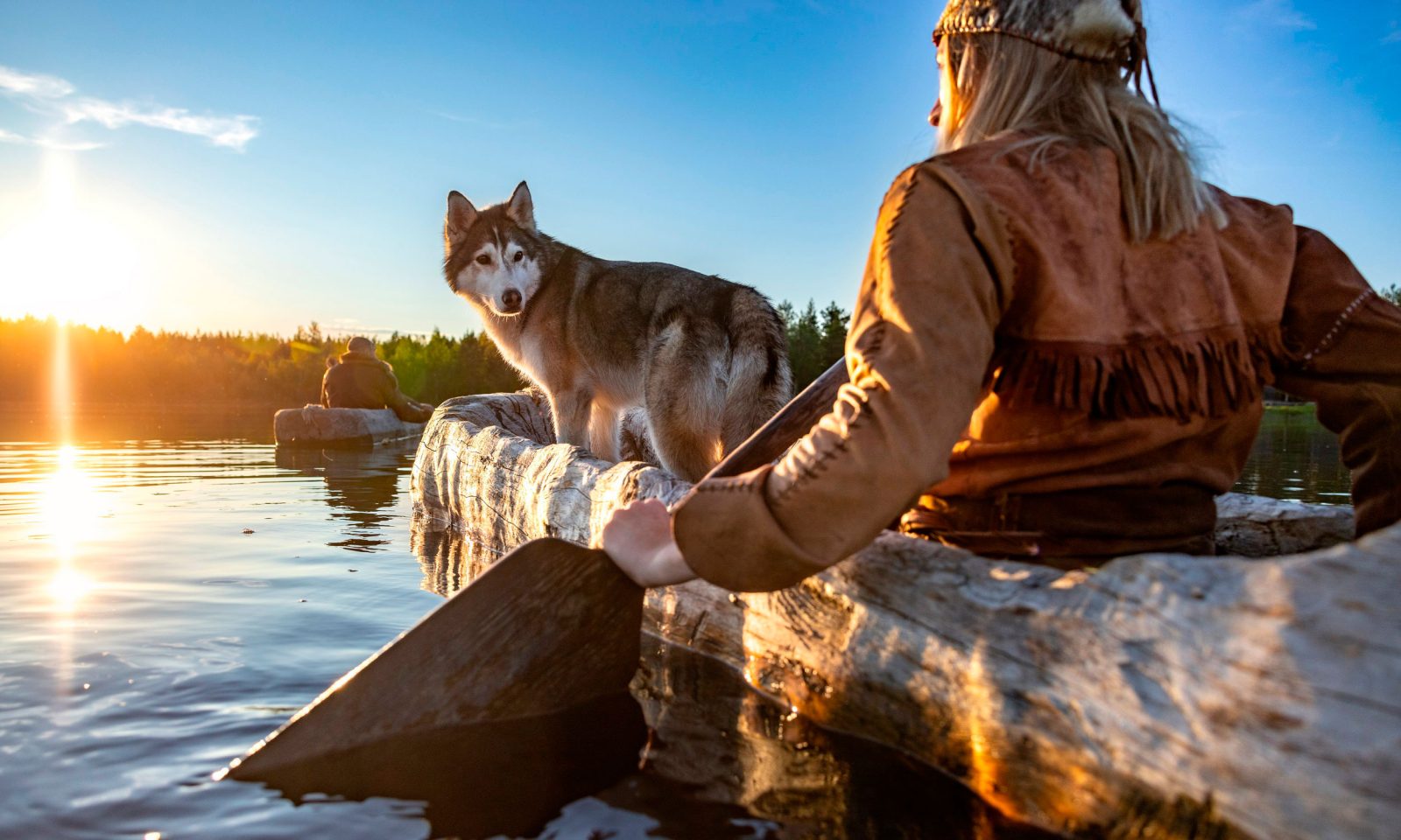 Nahka-asuun pukeutunut henkilö ja koira puisessa ruuhessa vesillä auringon laskiessa