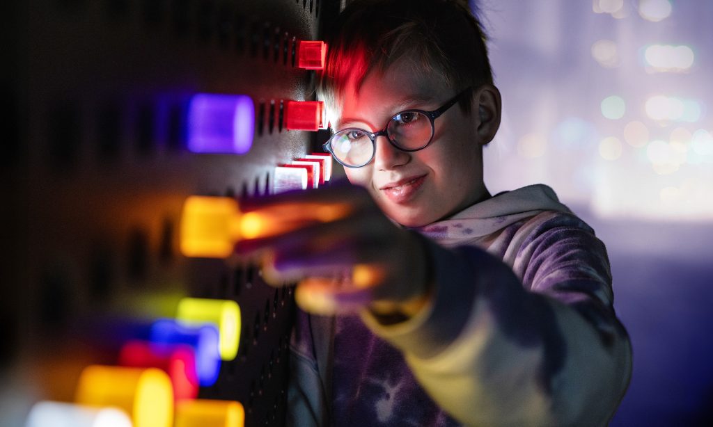 A child looks closely at a large board with colorful, illuminated pegs. The surroundings are dim and there is a soft, colorful bokeh effect in the background.