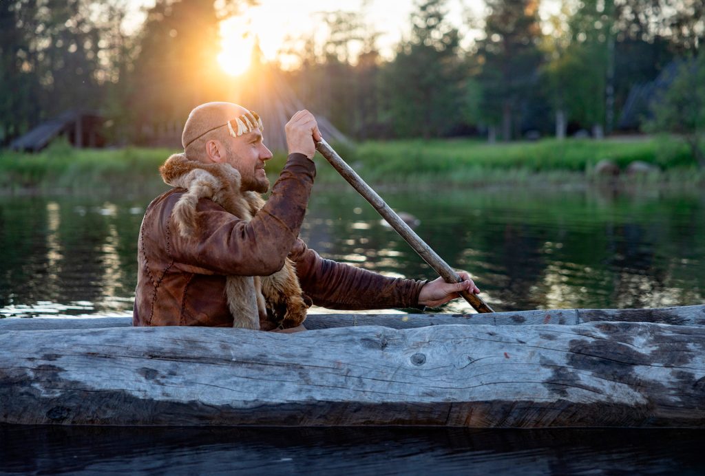 Nahka-asuun pukeutunut henkilö meloo puusta veistetyllä ruuhella, taustalla paistaa auringonlasku.