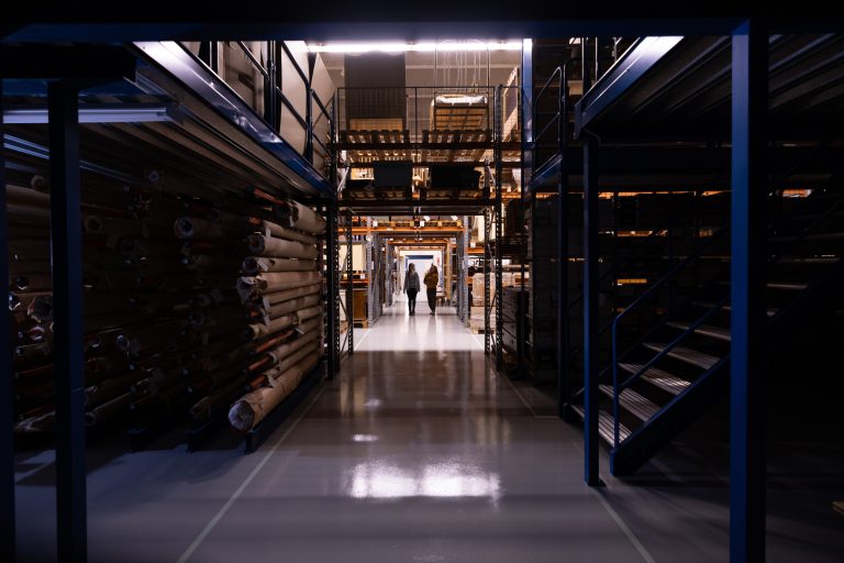 Varastokäytävä, molemmilla puolilla aseiden metrien kokoisia hyllyjä täynnä museon kokoelmia.