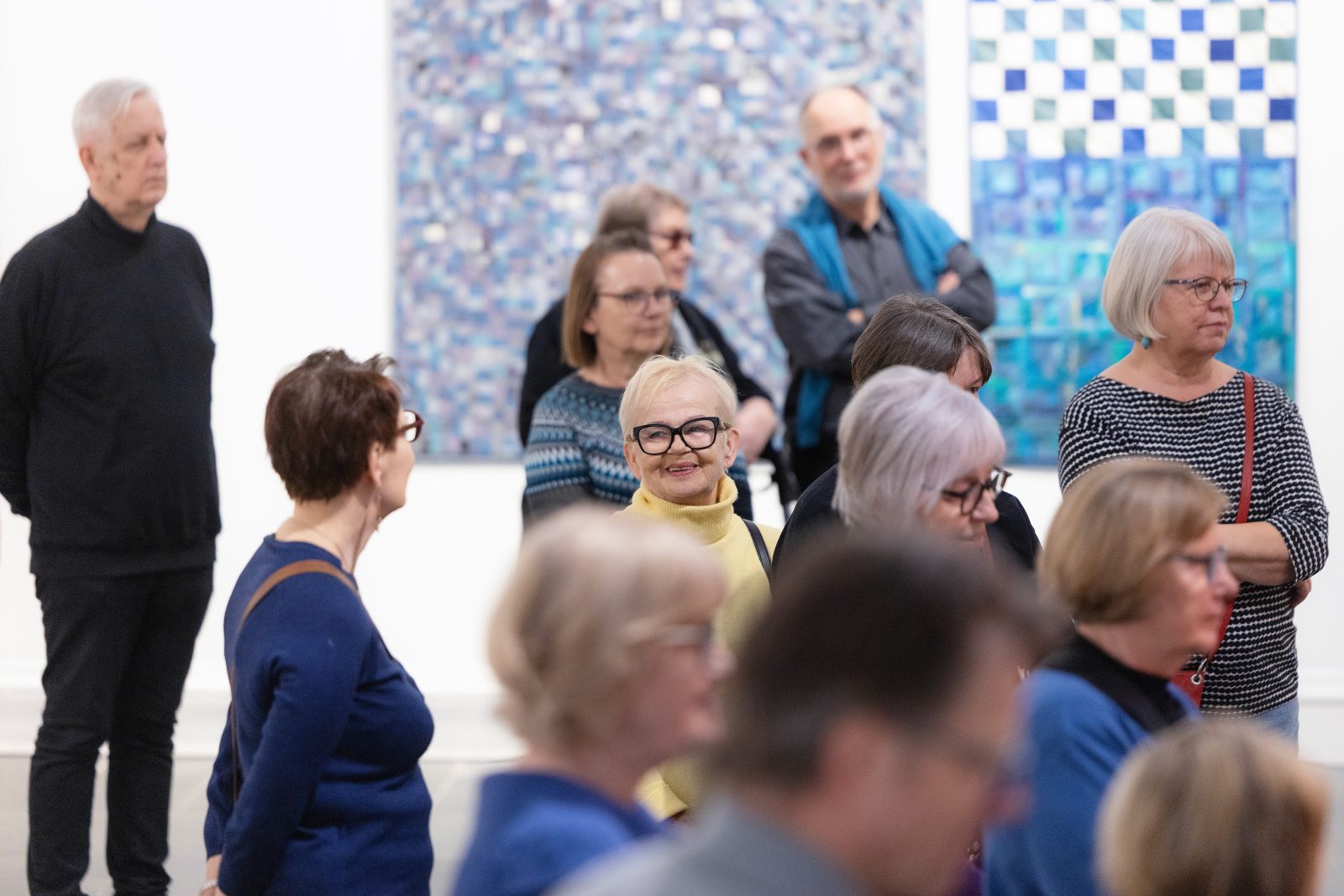 Useita henkilöitä seuraamassa näyttelyesittelyä, kuvan keskellä hymyilevä henkilö, jonka kasvot ovat kameraan päin. Taustalla kankaasta tehtyjä taideteoksia.