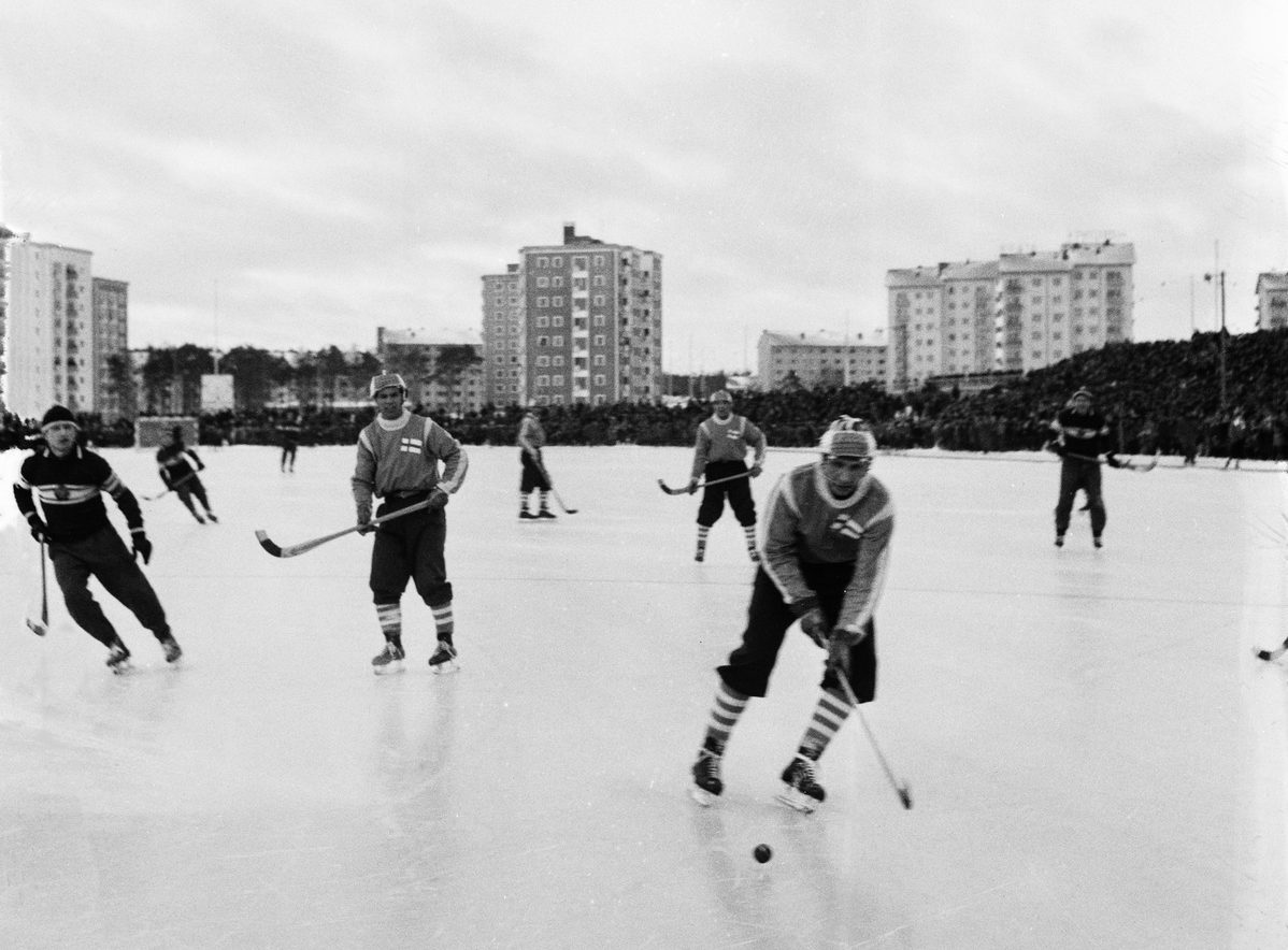 Jääpallon pelaajia jäällä, taustalla kerrostaloja.