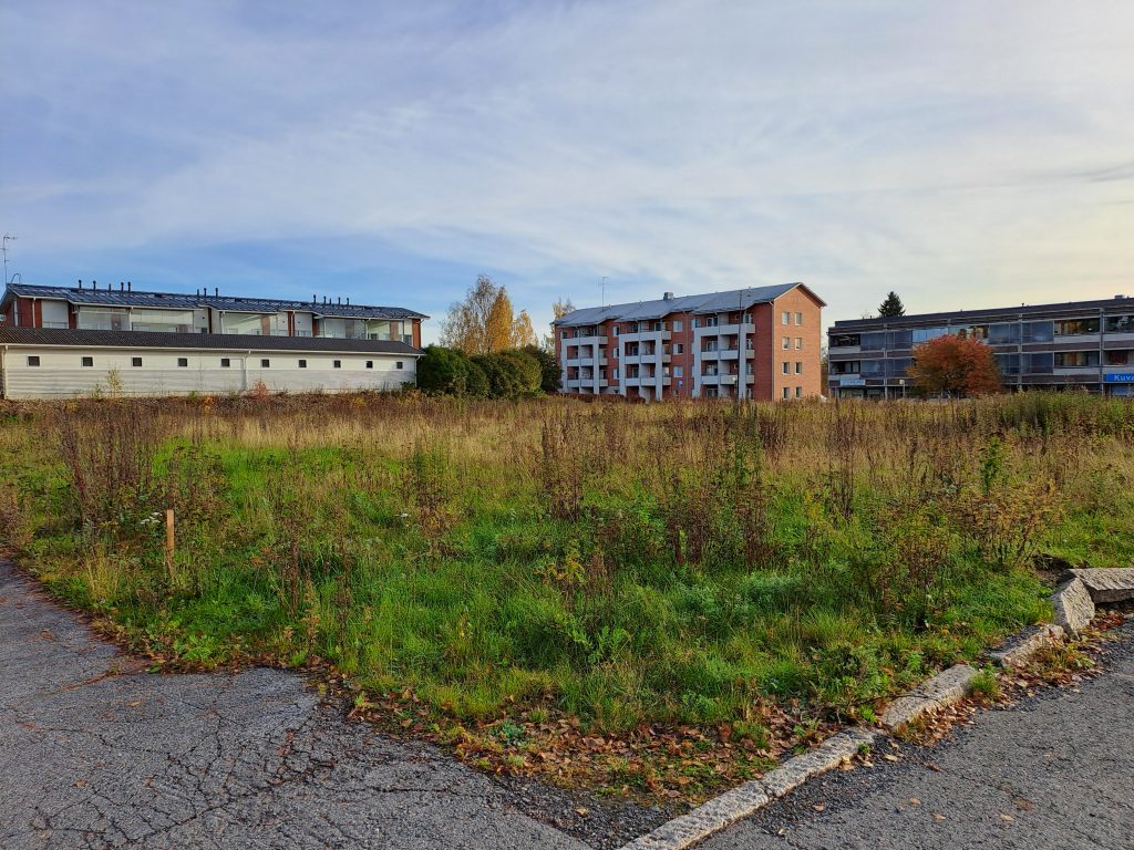 Tyhjä ruohikkoalue, taustalla matalia kerrostaloja.